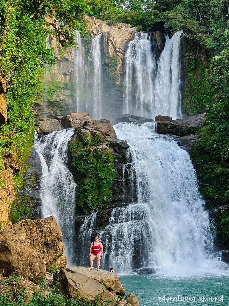 A woman standing on a boulder in front of a tall waterfall in Costa Rica.